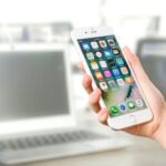 Close-up view of a smartphone displaying apps, held by a hand, with a blurred laptop in the background.